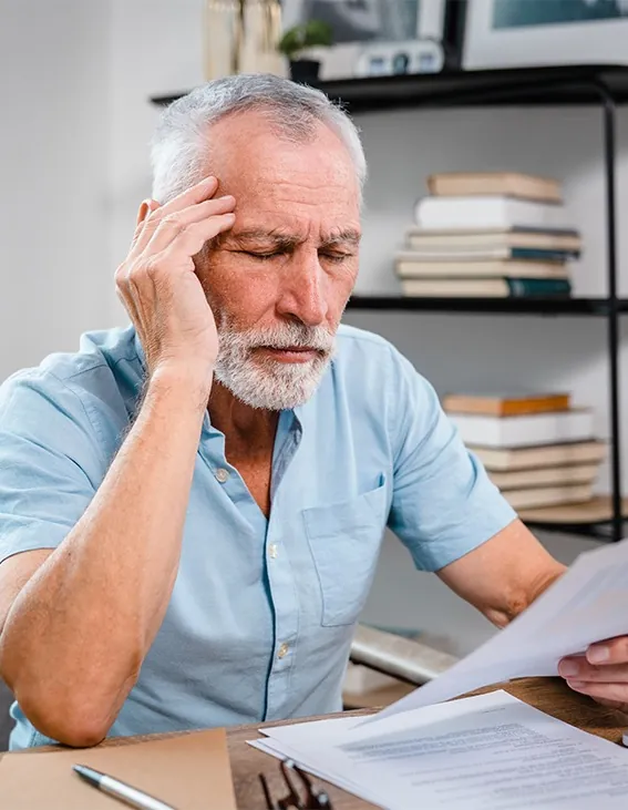 Older man with a hand on his temple looking stressed over papers at a desk