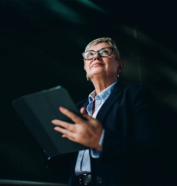 Older woman with glasses and a blazer holding a tablet, looking upward